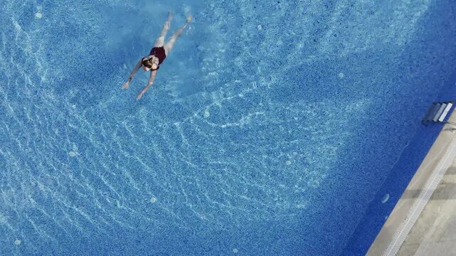 Drone top-down view of a young woman swimming in a crystal clear blue pool at a modern residential complex in Mahmutlar. Sunlit summer day scene with luxury amenities, loungers, umbrellas, and green l