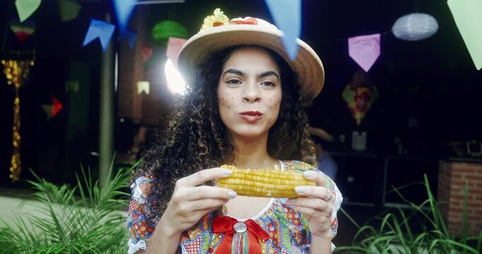 A cheerful Brazilian woman in traditional caipira attire and a straw hat enjoys eating a delicious grilled corn on the cob during a vibrant Festa Junina celebration with colorful decorations