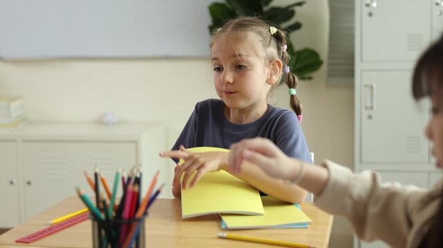 A young girl with pigtails is sitting at a desk, flipping through a notebook in a classroom setting with pencils and books around.