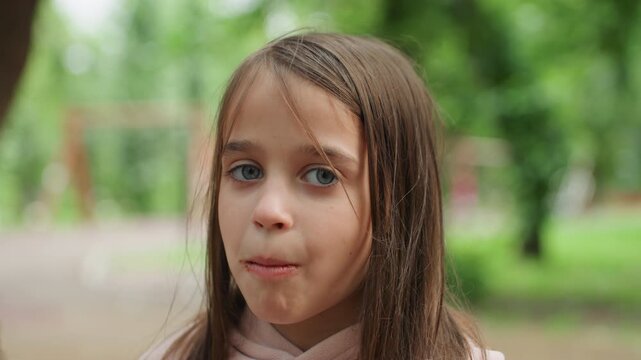 Child enjoying snack. Young girl with cookie near park. Child in hoodie savoring treat by playground trees. Young girl wearing hoodie chewing cookie represent in park setting during summer