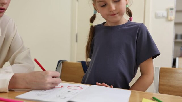 A young girl with braided hair is focused on drawing with a red colored pencil on a sheet of paper at a wooden table indoors.