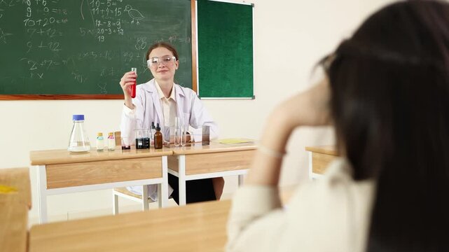 A female teacher wearing safety goggles demonstrates a chemistry experiment with test tubes in a classroom with a chalkboard in the background.