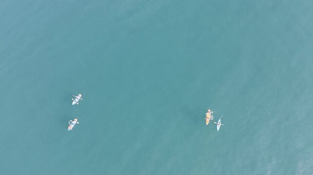Vertical top-down drone footage of people paddling in kayaks and on stand-up paddleboards (SUP) in crystal clear turquoise water. The shot captures the bright colors of the boats against the transpare