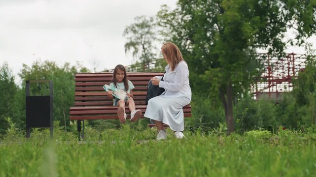 White mom and daughter sitting bench, mother offers snack and gentle advice while child fidgets with hands, warm interaction under trees, peaceful afternoon vibe, family bond and patient guidance,