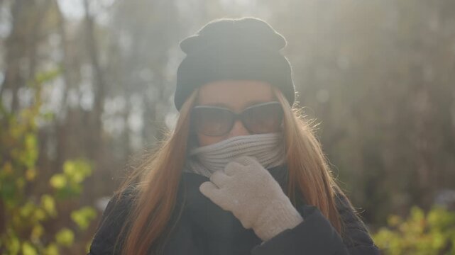 Closeup red haired white woman smiling in sunlight, wearing sunglasses, knit scarf and gloves, playful expression with coy hand gesture, backpack visible in background, warm golden backlight, dust