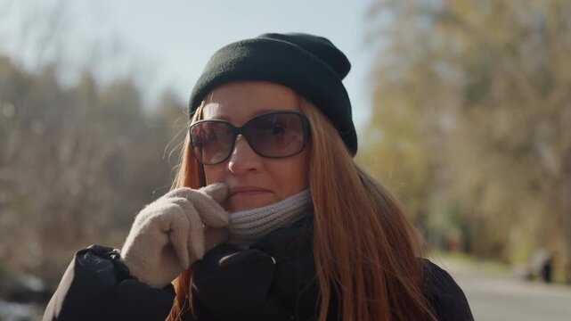 White woman outdoors adjusting scarf mask, red hair with beanie and sunglasses gloved hand checks surroundings, wary expression, autumn park pathway, soft sunlight and shallow depth, implied snatcher