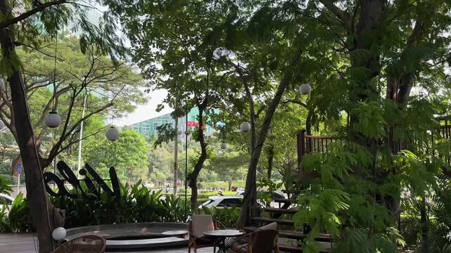 A steady, high-quality video shot from inside a lush, tranquil garden cafe. The footage showcases a cozy seating area on a rustic wooden terrace deck, featuring comfortable modern chairs.