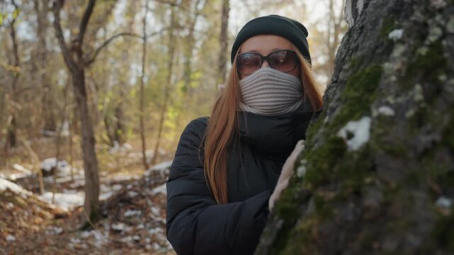 Woman snatcher peeks around birch tree, surveying quiet trail with gloved hand on bark, layered winter clothes and sunglasses obscure identity, muted light through bare branches creates tense