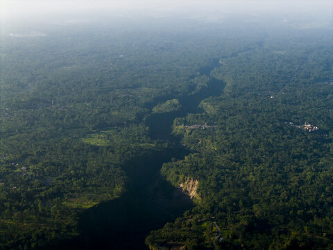 Aerial view of the deep Kali Gendol river valley surrounded by dense tropical forest and lush greenery under a hazy morning sky in Cangkringan, Special Region of Yogyakarta, Indonesia.
