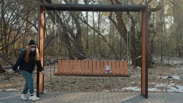 Park bench snatcher approaches unattended backpack on swing bench, crouches and grabs bag then walks away along leafstrewn path, cold winter light, beanie and coat, furtive motion captured by fixed