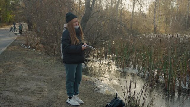 Woman standing by marsh holding controller, late autumn park pathway, puffy jacket and beanie, backpack on ground, reflective pond and reeds, distant pedestrians, quiet tense mood suggesting possible