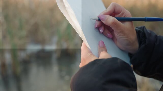 Close up hands writing by pond, field reporter taking brisk notes on small spiral notebook with pen, blurred reeds and cattails in warm backlight, attentive documentation for article or report,