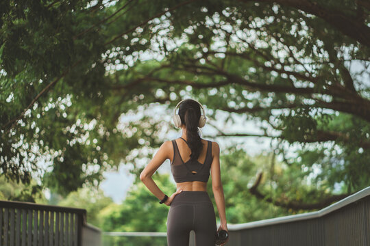 Back view of a fit young Asian woman in grey activewear with headphones and water bottle at a lush park. Preparing for outdoor workout or running at a city garden. Concept of health, wellness, and men