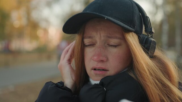 Closeup white redhead woman with headphones reacting to snatcher nearby in park autumn light, puffy black jacket, cap and scarf, anxious expression, hand to ear, searching gaze, candid street portrait