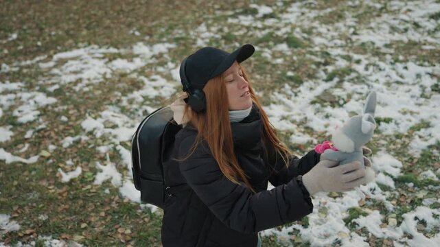 Caucasian woman holding plush bunny in snowy park, startled as unseen snatcher attempts grab wearing black coat, cap, headphones and scarf reactive hands reaching upward, tense expression, backpack