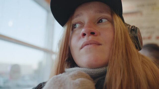 White redhead woman lowering scarf and revealing anxious eyes by window in cafe, headphones and cap still on flushed cheeks and watery gaze shift to regret and vulnerability, cinematic closeup