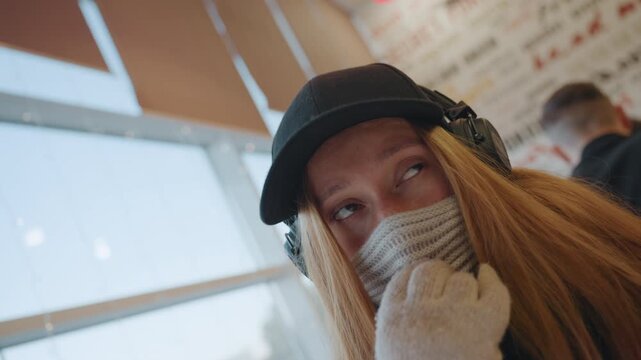Masked woman by window gazing at city skyline, headphones and cap, knit scarf and soft sunlit glow, transit lounge backdrop with distant passengers, introspective expression shifting between calm
