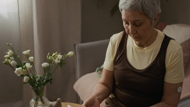 Tilt up shot of senior woman taking blood sample from finger then testing sugar level with glucometer sitting in living room during daily chronic condition management