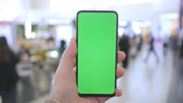 Hand holds smartphone with green screen. Background shows blurred indoor space. People walk in distance. Footage has three keyframes. No animals or plants shown
