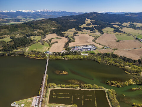 Aerial view of Oravska priehrada reservoir with a long bridge, surrounding green hills, and snow-capped Tatra mountains in the distance Namestovo, Zilinsky kraj, Slovakia.