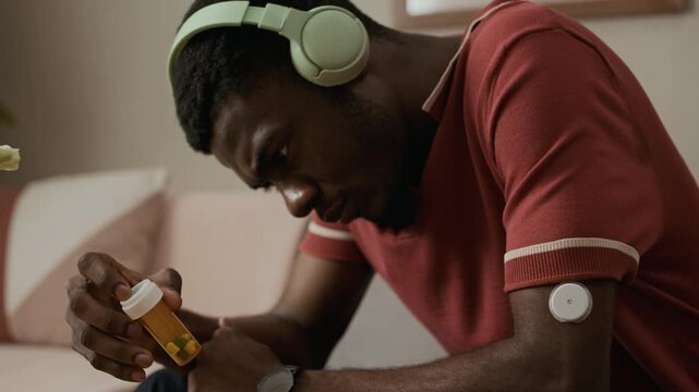 Close up shot of young adult African American man in headphones feeling concerned about taking diabetes pills while looking at prescription bottle in living room, tilt up camera movement