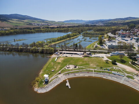 Aerial view of Oravska priehrada featuring a waterfront promenade with tiered stone seating, green park area, and rolling hills in Namestovo, Zilinsky kraj, Slovakia.