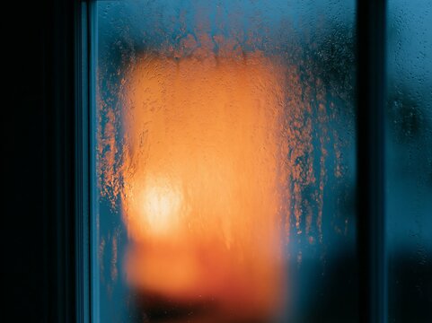 Close up of window with rain running down the glass, blurred warm light visible through the droplets, creating a cozy indoor atmosphere