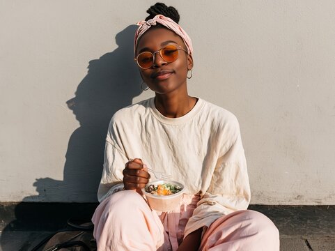 Young black woman wearing orange sunglasses and a headscarf sits against a wall, enjoying a bowl of food while dressed in a casual white top and pink pants