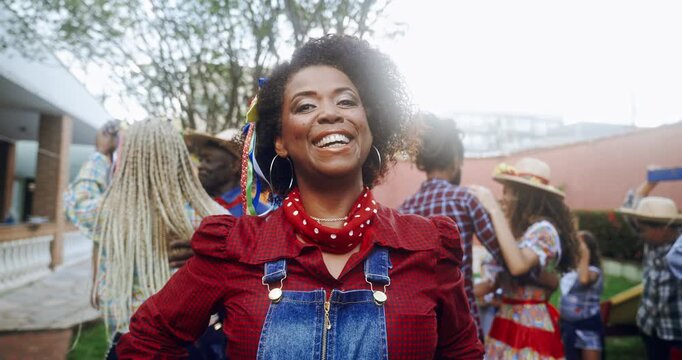 Joyful African American woman in traditional Festa Junina outfit smiling at the camera during a lively outdoor celebration with people dancing in the background on a sunny day in Brazil