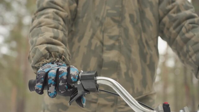 Tight shot atv handlebar and gloves, camouflaged rider testing throttle and brake, adjusting grip on chrome bars, pine forest backdrop, calm focus and readiness for offroad adventure
