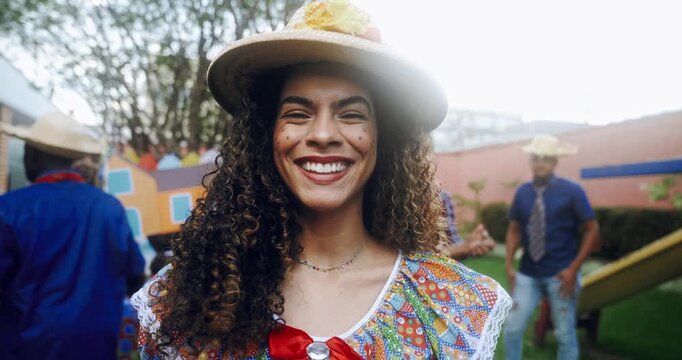 A beautiful young woman with curly hair smiles radiantly while wearing a traditional straw hat and colorful floral dress during a festive Brazilian Festa Junina celebration in an outdoor setting