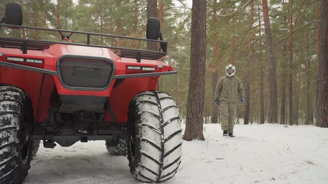 Rider approaches red atv in snow, puts gloved hand on front wheel, inspects tread and grille, scans surrounding pine forest, calm cautious mood, preparation for offroad extreme route