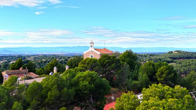 Small traditional stone chapel with a white bell tower surrounded by dense pine forest overlooking a distant mountain range. Stone church building with bell tower among green trees.