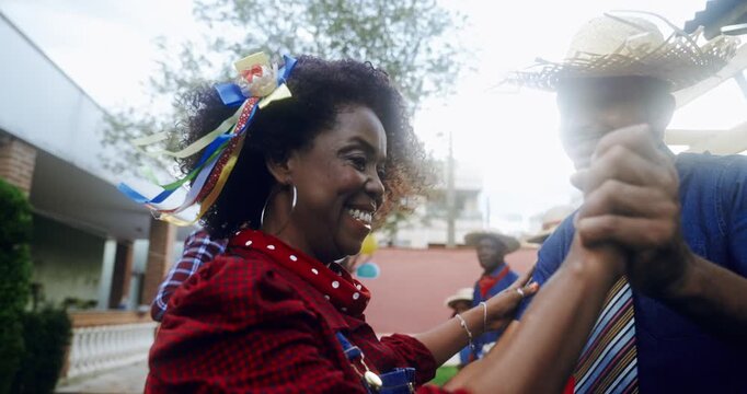 Happy group of diverse friends celebrating Festa Junina in Brazil, dancing and laughing outdoors in traditional costumes like plaid shirts and straw hats during a sunny day celebration