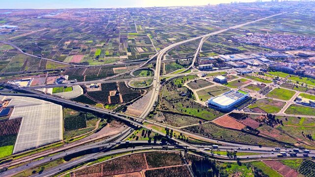 Large road junction with ramps and bridges integrated into a rural farming landscape. Complex highway interchange surrounded by agricultural plantations.