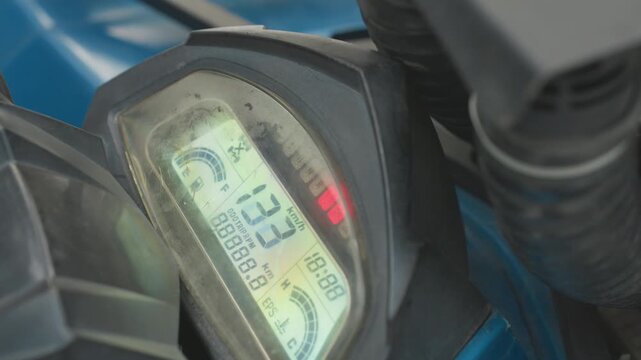 Atv dashboard display showing digital readouts, scratched plastic cover, illuminated lcd odometer and rpm numbers, red warning led glowing, blue bodywork and handlebar cables in frame, macro closeup