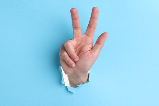 Man showing number three with fingers through hole in light blue paper, closeup