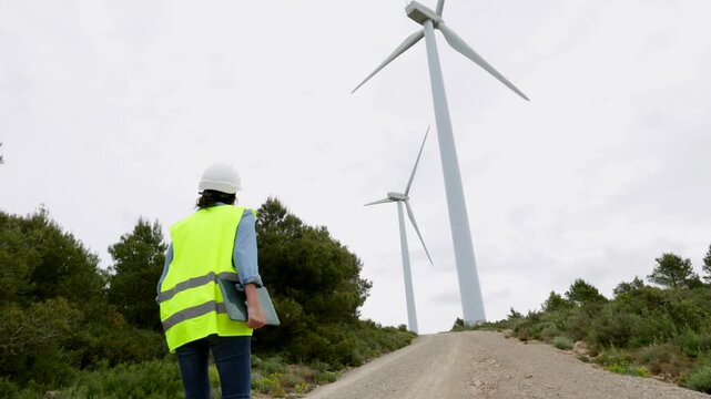 Female engineer walking toward wind turbines on a wind farm, inspecting renewable energy production. Professional worker with safety vest and hard hat on site