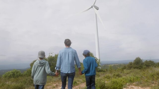 Mother and two sons holding hands walking towards a wind turbine, showing renewable energy to the next generation and promoting a sustainable future with clean electricity
