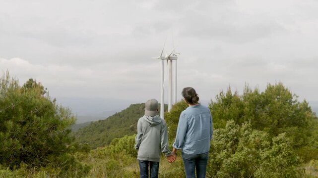 Young mother holds her son&rsquo;s hand as he points to wind turbines on a green mountain, learning about renewable energy, conservation, and a sustainable future together
