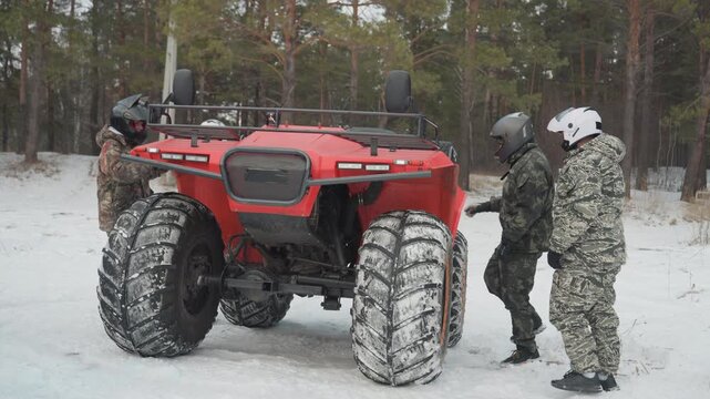 Crew inspects red quad in snowy pine forest before departure. Mechanic checks engine, tightens bolts, team mounts vehicle and secures gear, helmets on, hands on wrench, focused prep and rugged