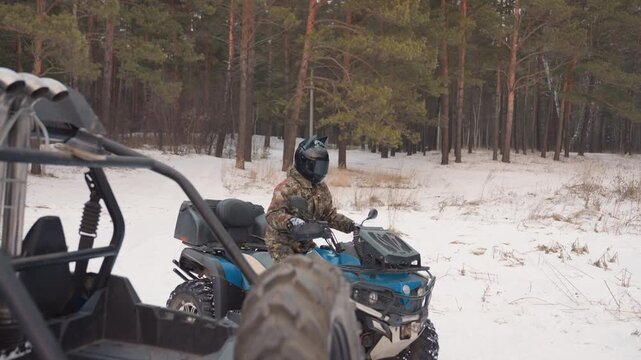 Rider inspects atv in snow near pine trees, walking around blue quad checking tire, winch and engine, adjusting helmet and straps, methodical maintenance before extreme trail ride
