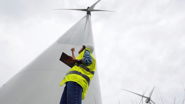 Engineer in hard hat and hi-vis vest inspects wind turbine outdoors, using a tablet to monitor performance and ensure sustainable, safe renewable energy production