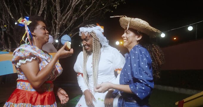Three happy Brazilian women in traditional Festa Junina costumes celebrate the June festival at night, laughing and eating typical snacks while wearing colorful caipira and bridal outfits