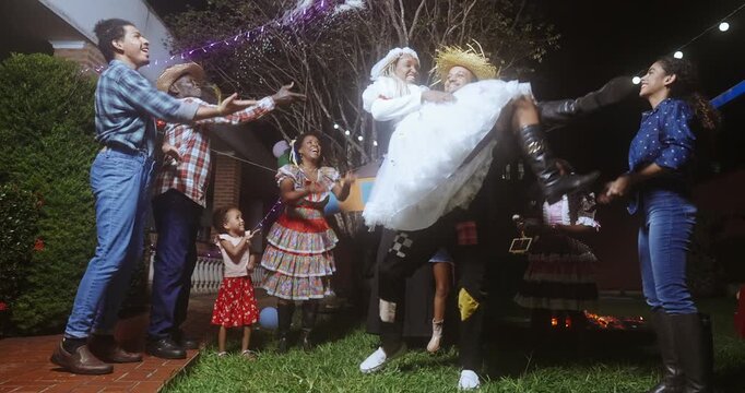 People celebrating a traditional Brazilian Festa Junina at night, featuring a mock wedding ceremony where a man lifts a bride in a white dress while guests in caipira costumes cheer and dance