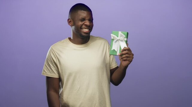 Young black man in beige tshirt holding a green wrapped gift with white bow, smiling and holding it up in a studio purple backdrop; joy celebration gratitude.