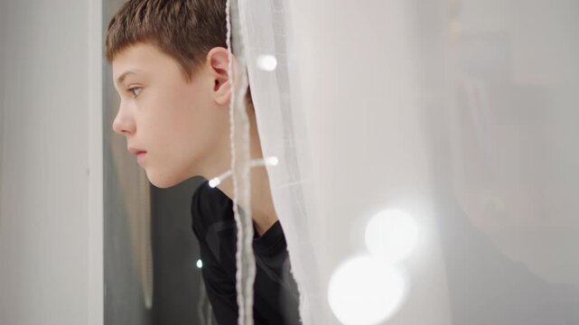 Grandchild looks through sheer fabric at sparkling nighttime illumination with hope. Younger family member observes luminous patterns beyond window with tender optimism and calm resolve