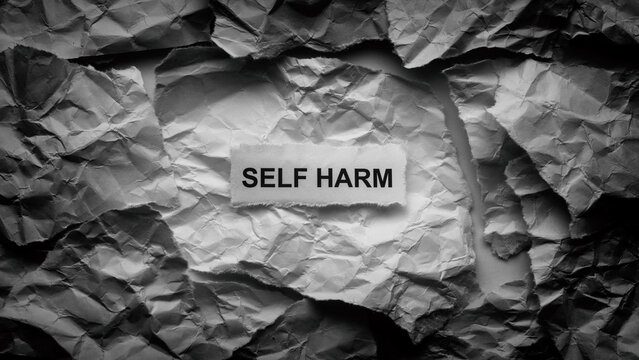 A torn piece of paper with the words Self harm on it lying on a background made of chaotic crumpled paper textures. Black and white. Close up.