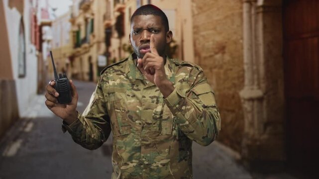 Young soldier in camouflage holding a radio, finger to lips for silence in a narrow stone street alley during urban patrol; caution vigilance duty.