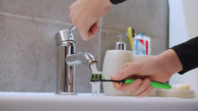 Closeup chrome faucet pouring water over toothbrush in modern bathroom sink, hand steadying green toothbrush, ceramic holders with toothpaste and soap, soft natural light and neutral tile background,